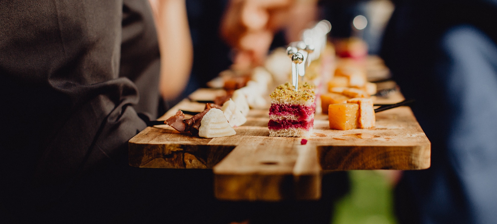 Un serveur présente un plateau rustique de bouchées salées, dont un canapé à la betterave et pistache, du fromage et de la charcuterie.